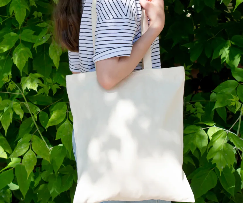 Person carrying a blank canvas tote bag in front of green foliage at Georgette's Grounds & Gifts, highlighting a Canvas Tote Bag Decorating Event.