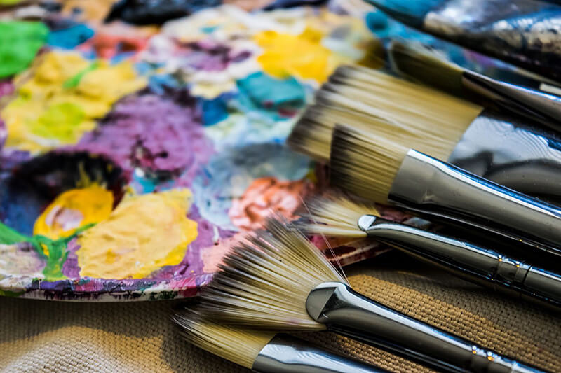A close-up of several paintbrushes lying next to an artist’s palette covered in bright, multicolored paint splotches, including yellows, purples, blues, and greens.