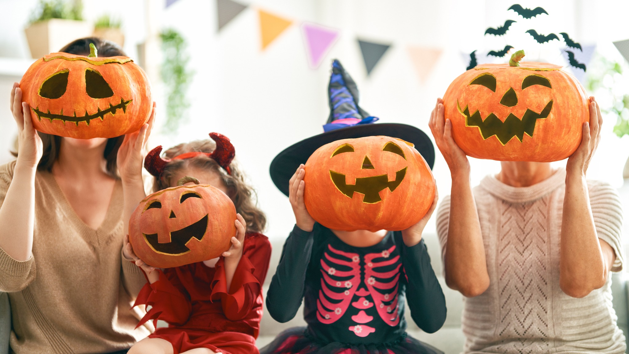 A family dressed in Halloween costumes holds carved jack-o’-lantern pumpkins in front of their faces. The group includes two adults and two children in festive outfits, one in a devil costume and another in a skeleton witch costume. Colorful pennant banners hang in the background