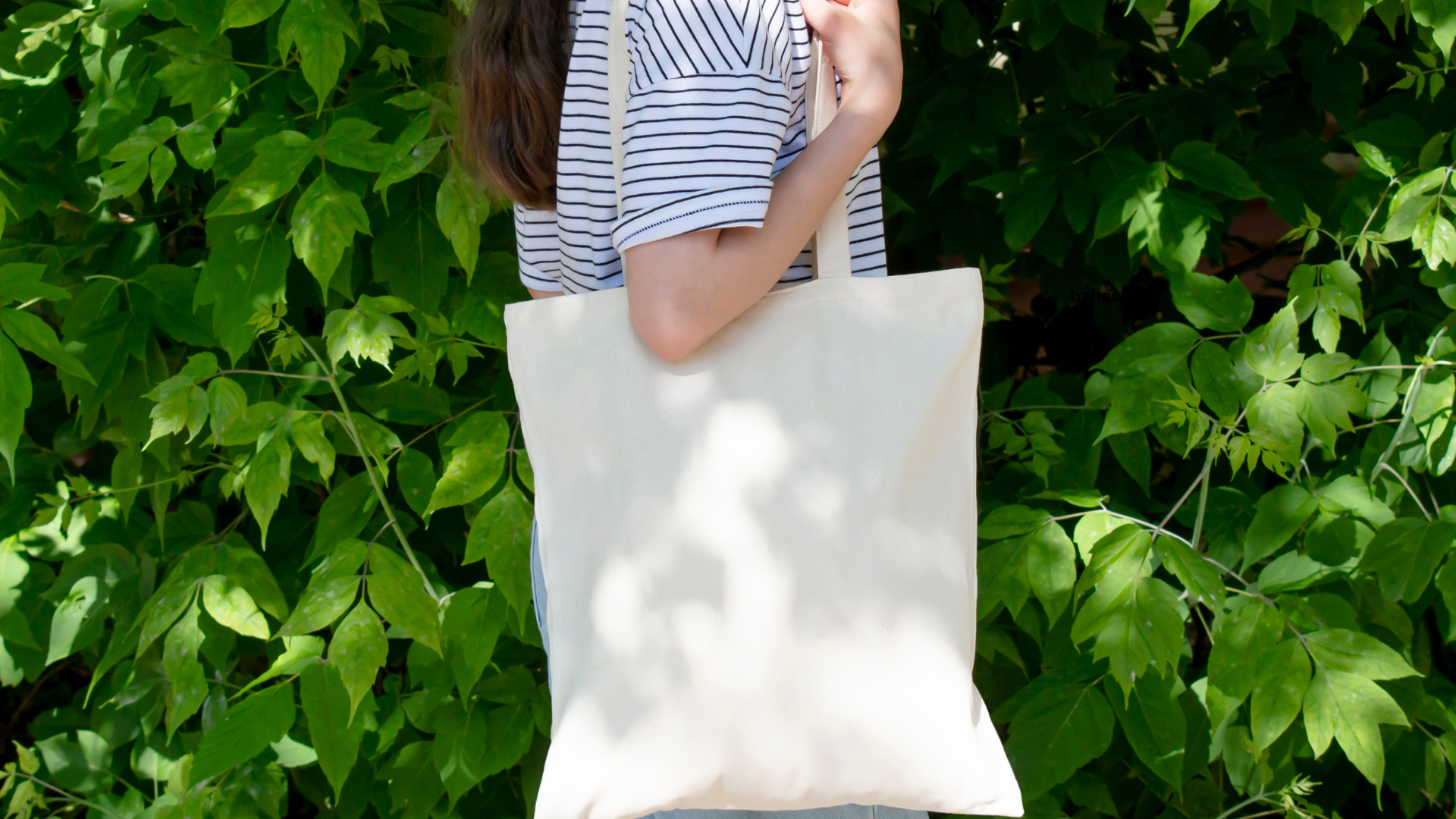 Person carrying a blank canvas tote bag in front of green foliage at Georgette's Grounds & Gifts, highlighting a Canvas Tote Bag Decorating Event.