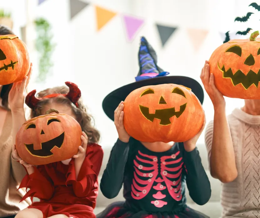 A family dressed in Halloween costumes holds carved jack-o’-lantern pumpkins in front of their faces. The group includes two adults and two children in festive outfits, one in a devil costume and another in a skeleton witch costume. Colorful pennant banners hang in the background