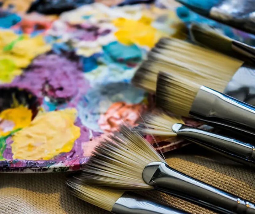 A close-up of several paintbrushes lying next to an artist’s palette covered in bright, multicolored paint splotches, including yellows, purples, blues, and greens.